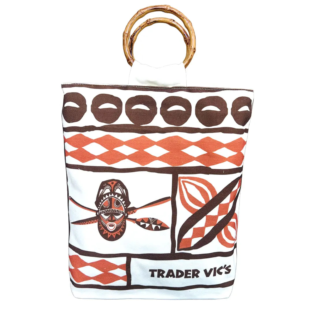 A white canvas tote bag with a brown and orange tapa pattern, featuring bamboo handle and the 'TRADER VIC'S' logo printed at the bottom.