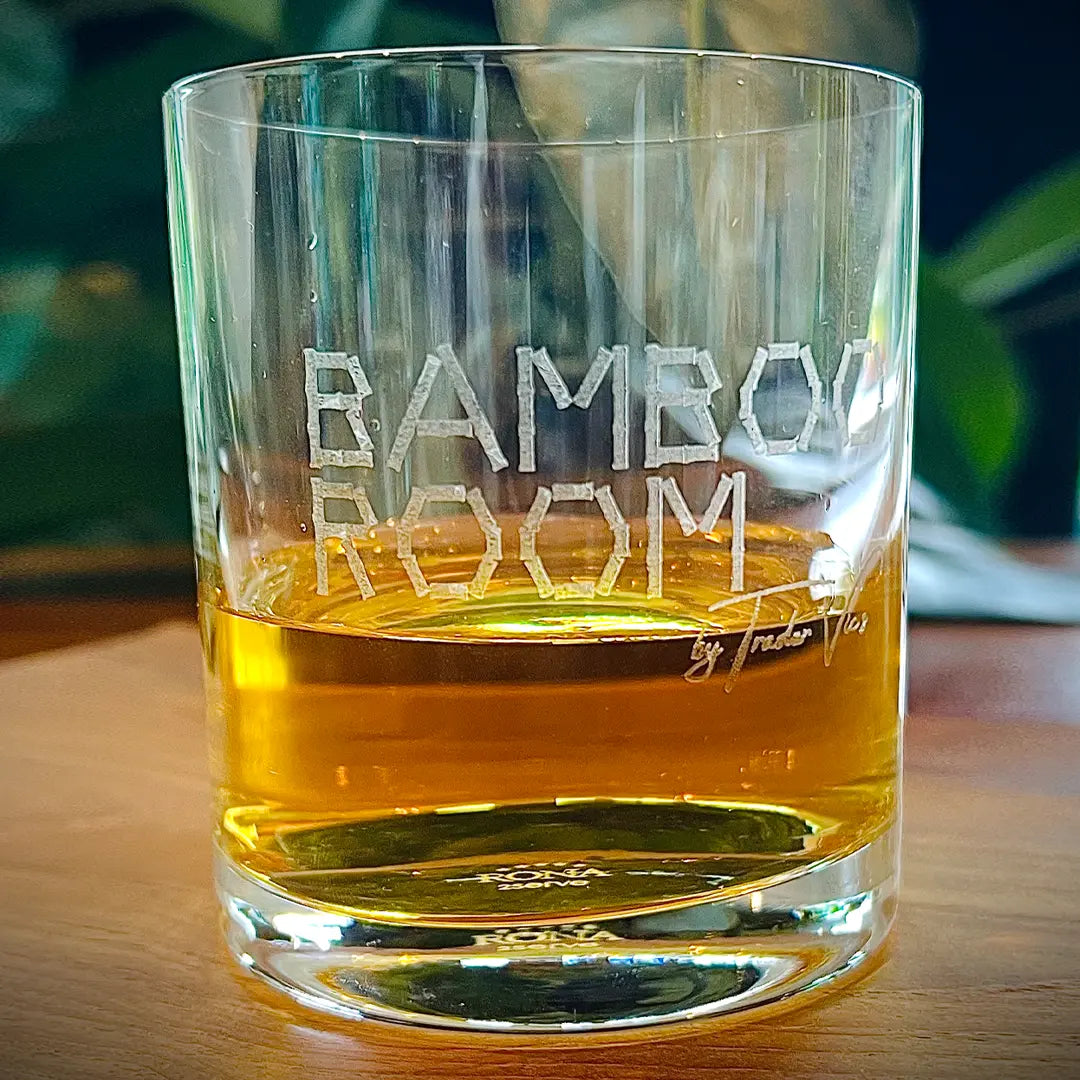 Glass filled with amber liquid on a wooden surface, with 'Bamboo Room' engraved on it.