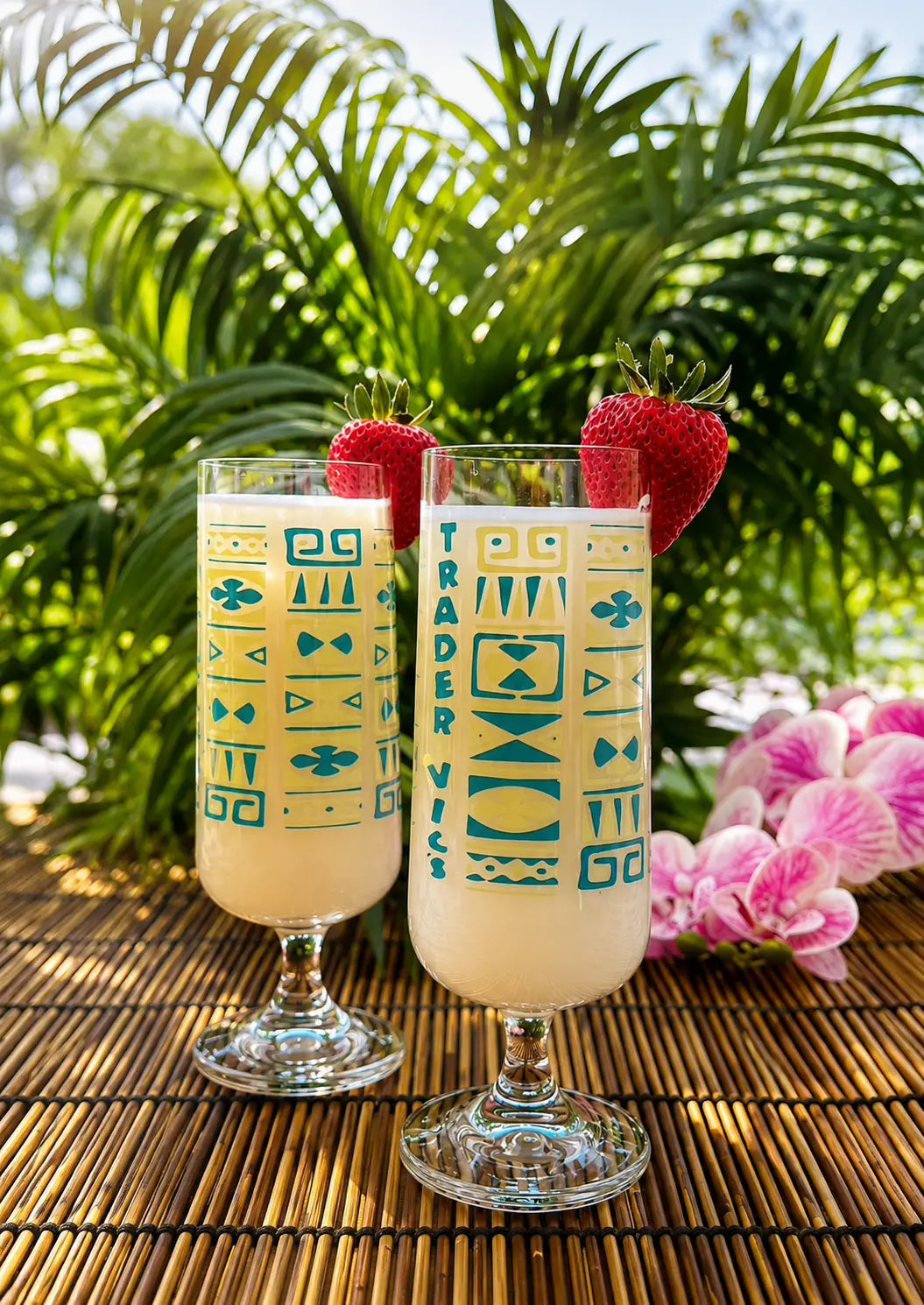 Two tall clear glasses filled with a creamy pale yellow beverage, decorated with teal geometric patterns and the vertical text 'TRADER VTS', sit on a woven bamboo mat outdoors with palm leaves and pink flowers in the background. Each glass is garnished with a strawberry.