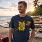 Man wearing a navy blue t-shirt with a yellow graphic design on a beach with boats and sunset in the background