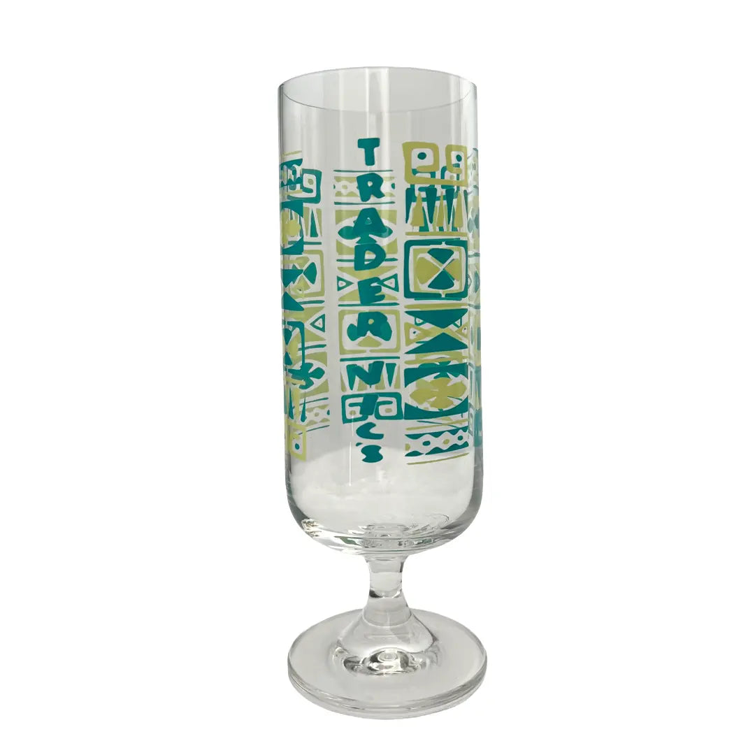 A clear glass goblet with a stylized teal and yellow-green geometric graphic pattern featuring the word 'TRADE' vertically, set against a plain white background.