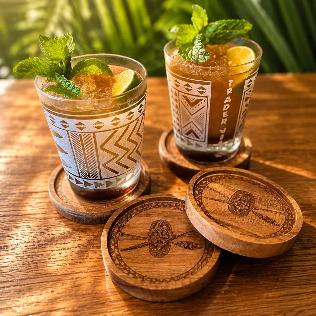 Two patterned drinking glasses filled with iced cocktails sit on wooden coasters on a wooden table, with two matching coasters in front and green foliage blurred in the background.
