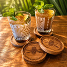 Two patterned drinking glasses filled with iced cocktails sit on wooden coasters on a wooden table, with two matching coasters in front and green foliage blurred in the background.