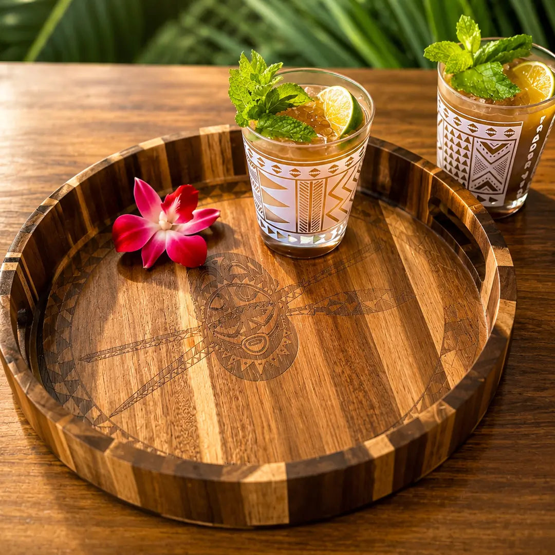A round wooden serving tray with engraved tribal-style patterns and striped edges, styled on a wooden table with two patterned drinking glasses containing iced drinks, mint, lime, and a pink flower.