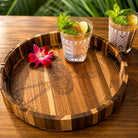A round wooden serving tray with engraved tribal-style patterns and striped edges, styled on a wooden table with two patterned drinking glasses containing iced drinks, mint, lime, and a pink flower.
